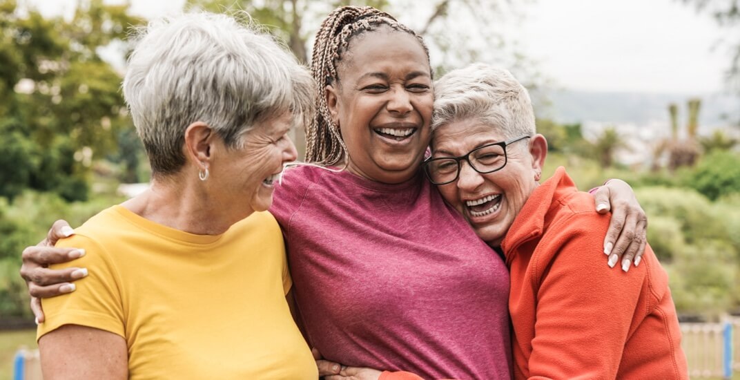 A group of three senior women laughing and hugging in a leafy park