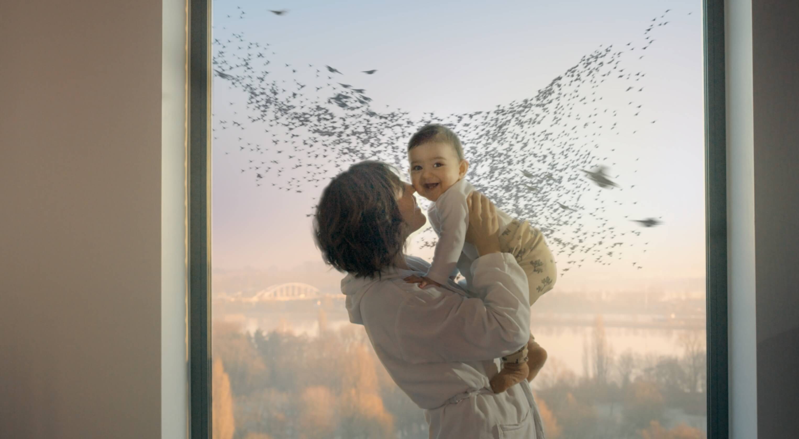 A woman embracing her baby. Through the window behind her, a murmuration of birds flies past