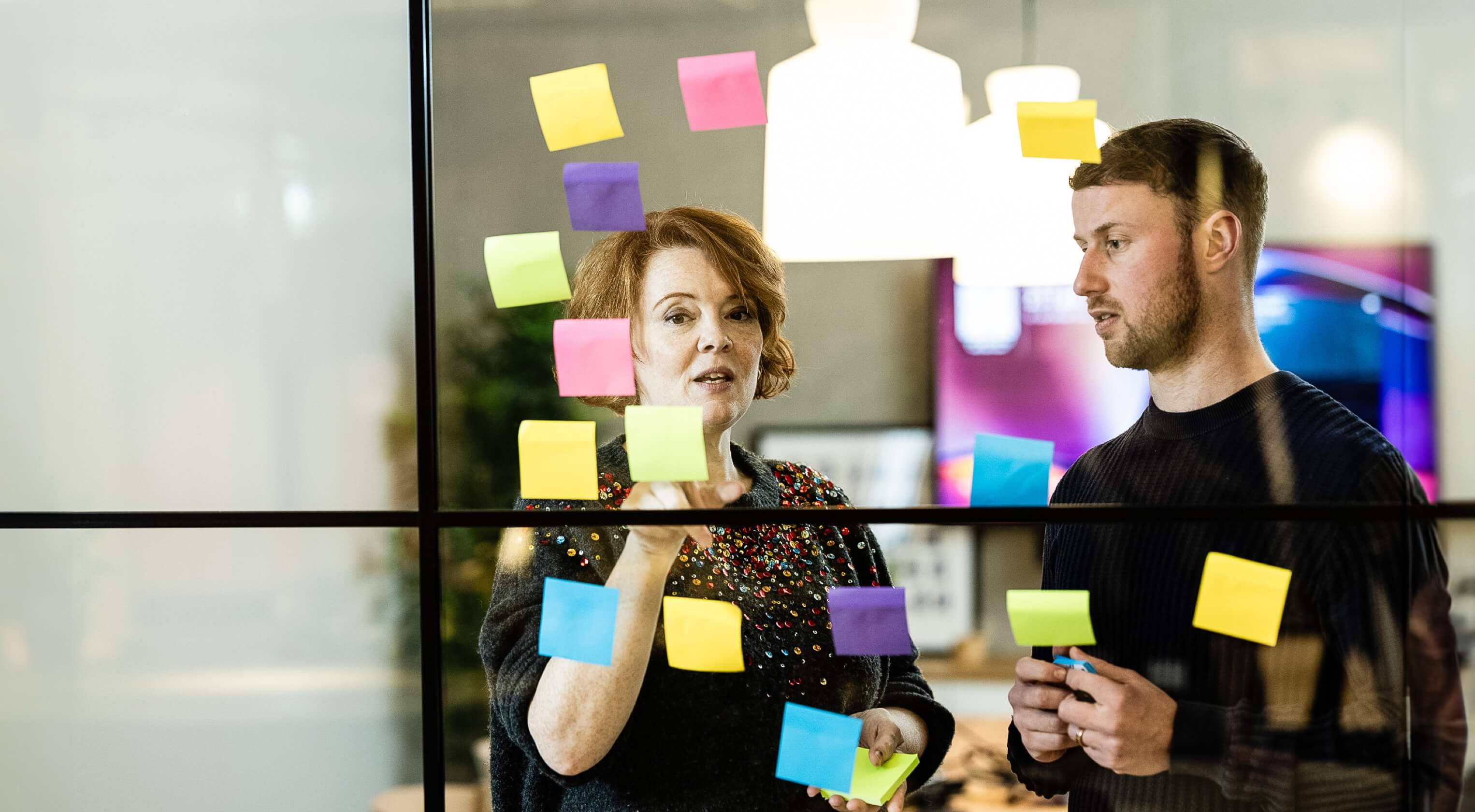 A man and woman talking whilst placing sticky notes on a glass partition