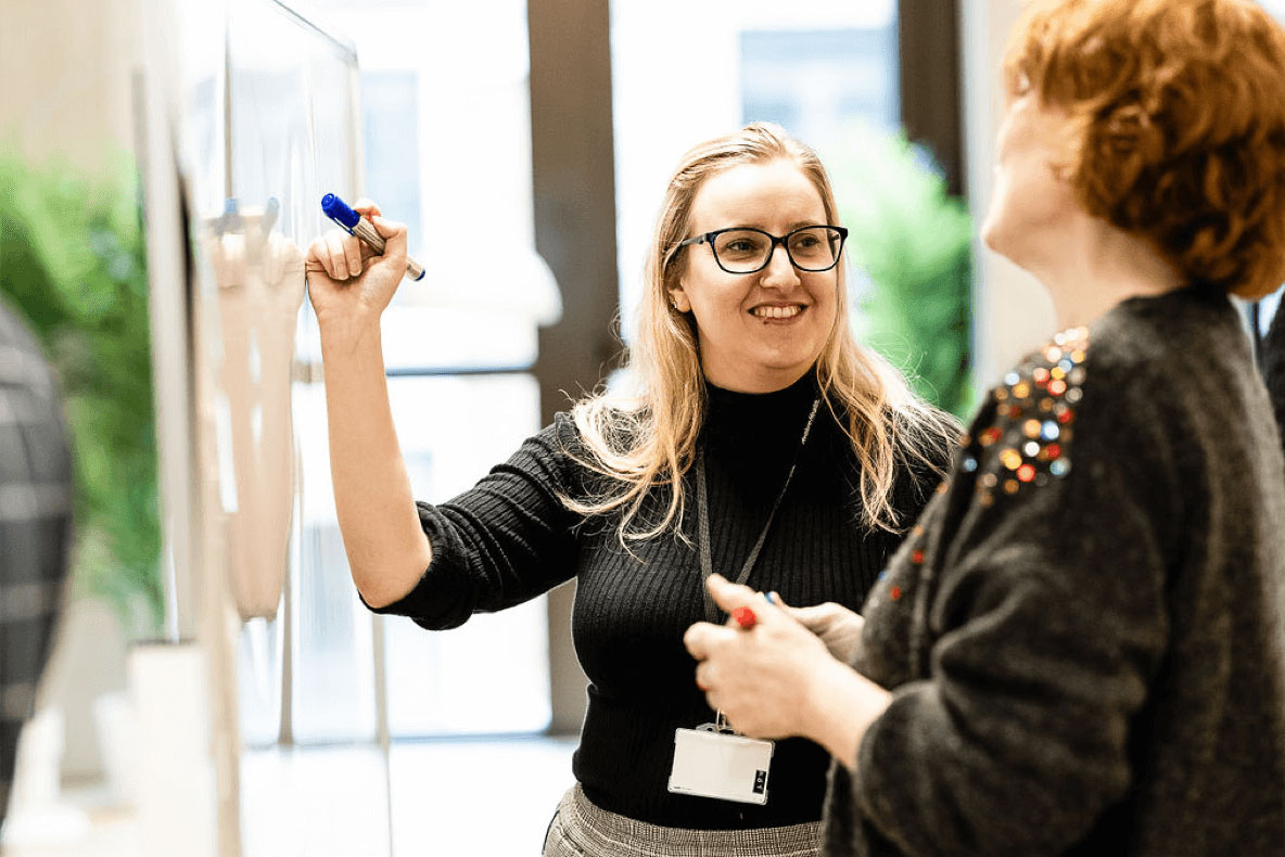 A woman writing on a white-board as another woman observes